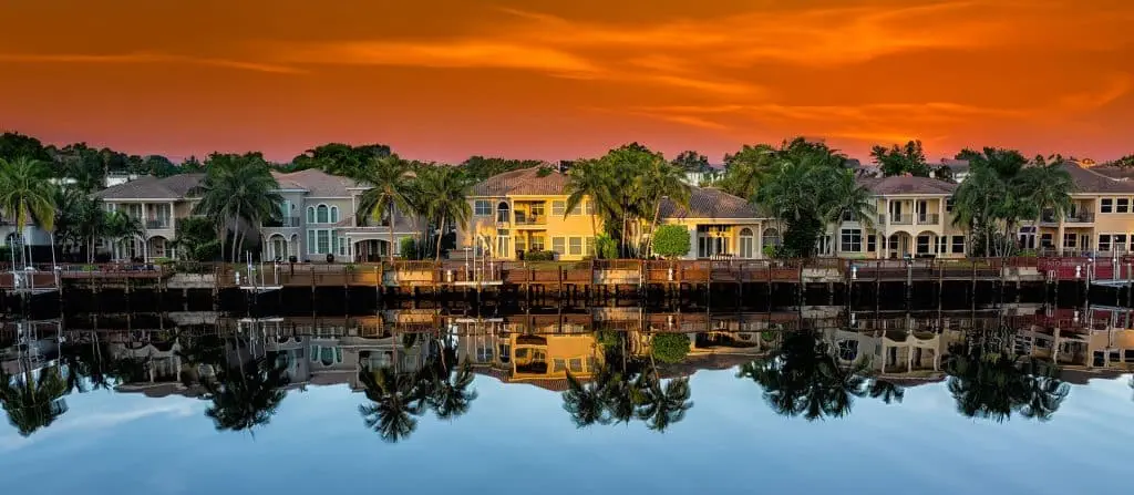 Row of waterfront houses with lush palm trees reflected in the calm water under a vibrant orange sunset sky.