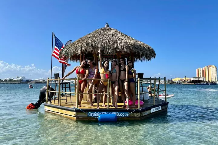 Group of people enjoying time on a cruisin tiki boat with a thatched roof, with buildings on the coastline in the background.
