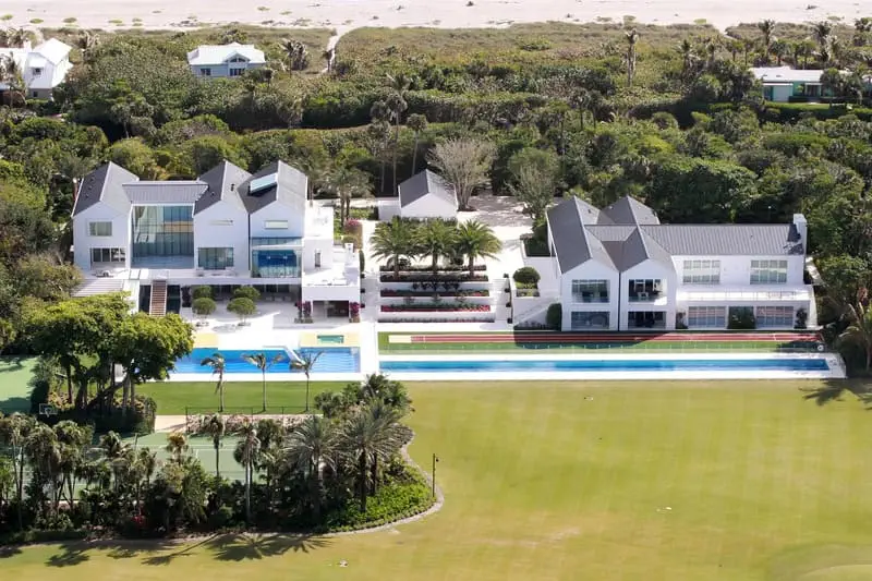 Aerial view of a Tiger Woods estate featuring two white buildings, a long swimming pool, tennis court, and landscaped gardens, surrounded by trees with a beach in the background.